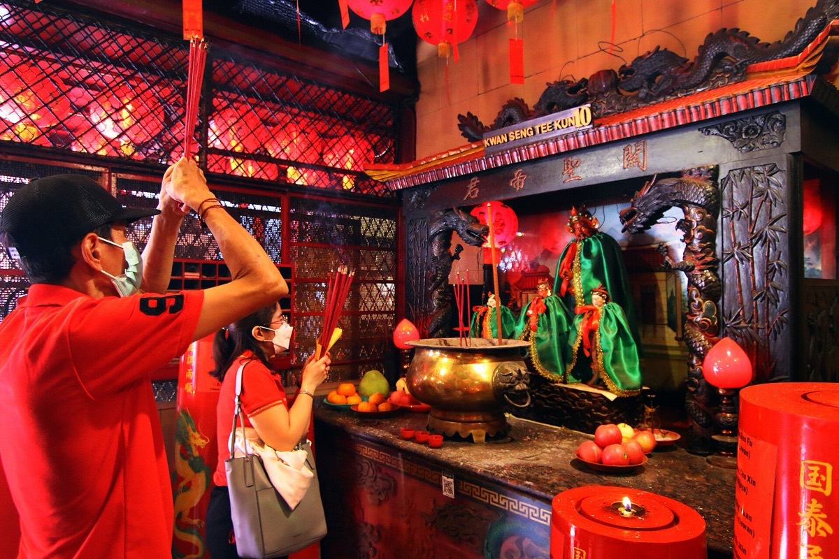 Lunar New Year prayer inside a Jakarta temple: two worshippers hold bundles of incense raised above their heads before a brass censer, with a lit red candle on a stand at the right.