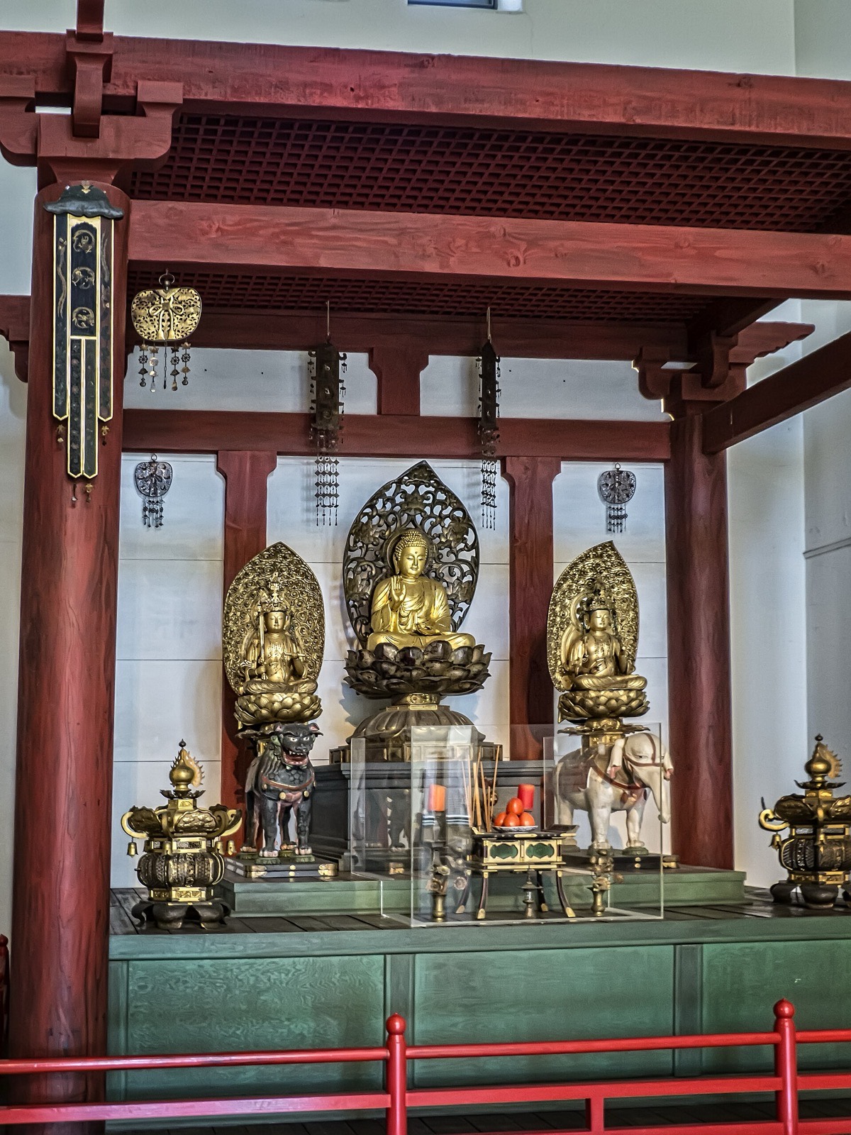 The inner sanctuary of a Shingon Buddhist temple: three gilt Buddhist figures on lotus pedestals under vermilion pillars, with a pair of orange pillar candles, incense sticks and fruit offerings on the table in front.